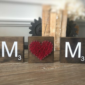 May include: Three wooden blocks with the letter "M" and the number "3" on each block. The middle block has a red heart made of string wrapped around nails.