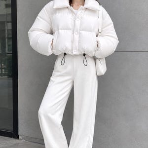 Op de afbeelding: Een wit kort gewatteerd jack met een brede kraag en drukknoopsluiting, gecombineerd met een bijpassende witte broek met wijde pijpen en witte sneakers. Een kleine witte schoudertas maakt de monochrome outfit compleet.