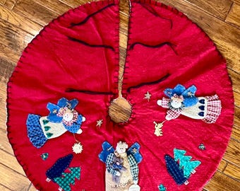 May include: A circular red felt Christmas tree skirt featuring three fabric angel appliqués with blue wings, plaid accents, and small green tree cutouts. The skirt has a central opening and black stitched edges with decorative gold stars.