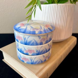 May include: A round, three-tiered porcelain trinket box featuring a delicate floral pattern of blue and pink irises with green leaves against a white background. The lid and each stackable section are finished with a thin, metallic gold rim.