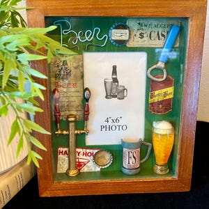 May include: A wooden shadow box frame with a green background featuring beer-themed decorations including a miniature tap handle, a bottle opener, bottle caps, a mug, and a glass of beer. The center contains a 4"x6" photo placeholder.