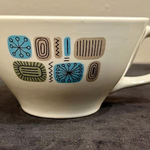 May include: A cream-colored ceramic cup featuring a repeating pattern of geometric shapes in shades of blue, olive green, and tan. The design includes starbursts, rectangles, and vertical lines, with a curved handle on the right side of the cup.