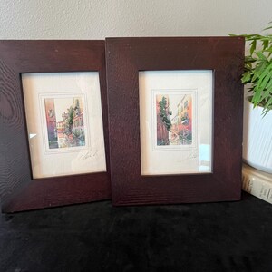 May include: Two framed watercolor paintings featuring narrow canal scenes with warm-toned buildings and greenery. Each artwork is set within a wide, dark brown wooden frame with a visible grain texture and a white mat board border surrounding the art.