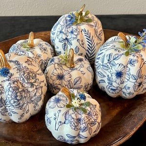 May include: A collection of six white decorative pumpkins featuring a blue floral pattern. Each pumpkin has a metallic gold stem and is accented with small artificial blue and white flowers and green leaves attached to the top of the stem.