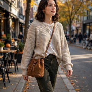 Puede incluir: Un cárdigan de punto acanalado beige sobre una camiseta blanca, combinado con pantalones verde oliva oscuro y un bolso tipo cubo de ante marrón con cierre de cordón. El conjunto se completa con botines marrones, creando un estilo otoñal.