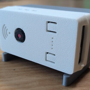 Könnte beinhalten: Ein rechteckiges elektronisches Gerät mit einer weißen Frontplatte und einem grauen Gehäuse. Die Vorderseite zeigt ein zentrales kreisförmiges Kameraobjektiv, ein WLAN-Signal-Symbol und eine vertikale Reihe von vier kleinen Leuchtanzeigen.