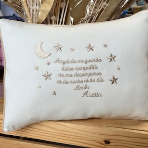 May include: A white pillow with a beige embroidered design of a crescent moon, stars, and a Spanish prayer. The prayer reads "Angel de mi guarda dulce compañía no me desampares ni de noche ni de día Amén Austin".