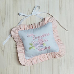 May include: A light blue pillow with pink ruffled trim and embroidered text that reads "Shh... Emmeline is sleeping" with a floral design.