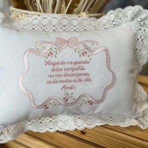 May include: White pillow with a lace trim and pink embroidered text that reads "Angel de mi guarda dulce compañía no me desampares ni de noche ni de día Amén".