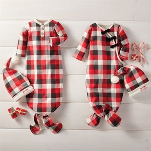May include: Two red and black plaid baby gowns with matching hats. The gowns have long sleeves and are tied at the bottom. The hats have pom-poms on top. The text "My First Christmas" is on the hats.