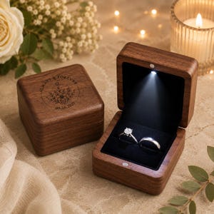 May include: Two square dark wood ring boxes with a hinged lid. One box is closed, featuring a laser-engraved floral design with the text Always & Forever and 08.28.2025. The open box contains two silver rings on a black velvet insert with an LED light.
