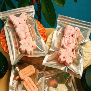 May include: Four clear resealable plastic pouches containing various shaped bath bombs. Two pouches hold pink, speckled floral and heart-shaped items. One pouch contains three light orange popsicle-shaped items. One pouch holds pale yellow cactus-shaped items.