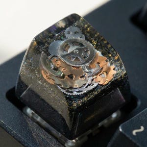Può includere: Un tasto meccanico quadrato realizzato in resina trasparente con base nera. L'interno è riempito con una collezione di ingranaggi e ruote dentate metalliche color argento e rame, sospesi nella resina insieme a sottili glitter dorati.