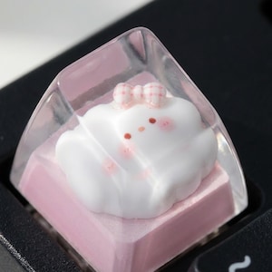 May include: A clear, square mechanical keyboard keycap featuring a sculpted white cloud figure with a small pink checkered bow on top. The cloud has tiny red dots for eyes and pink blush marks on its cheeks, set against a solid light pink base.