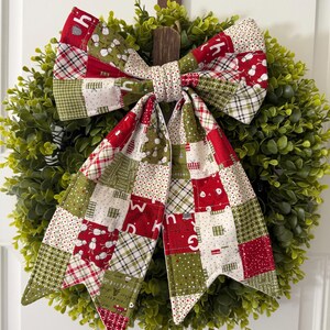 May include: A decorative fabric bow featuring a patchwork design of red, green, and white squares with holiday-themed patterns including snowmen, gifts, and plaid. The bow has two long, pointed tails and is centered on a green artificial boxwood wreath.