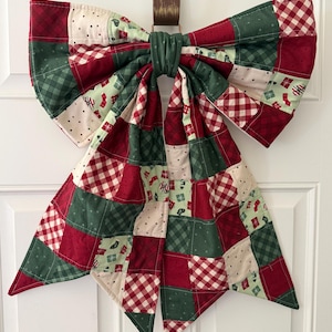 May include: A large fabric bow constructed from a patchwork of quilted squares in red, dark green, and cream. The fabric features patterns including gingham, polka dots, and small holiday-themed icons like stockings and gift boxes.