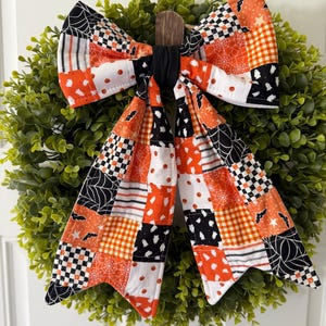 Könnte beinhalten: Eine dekorative Stoffschleife mit einem Patchwork-Muster aus orangefarbenen, schwarzen und weißen Halloween-Motiven, darunter Geister, Fledermäuse, Spinnennetze, Kürbisse und Karomuster, in der Mitte mit einem schwarzen Stoffknoten versehen.