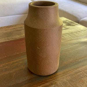 May include: A cylindrical vase with a matte, earthy brown finish and a slightly tapered neck. The surface has a fine, granular texture throughout, and the opening at the top is circular with a smooth, thick rim, creating a simple, functional form.