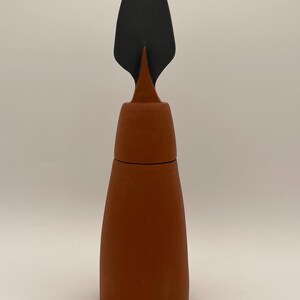 May include: A tall, cylindrical terracotta-colored ceramic vessel with a matte finish. The object features a distinct, tapered top section topped with a flat, spade-shaped black handle or lid component that extends vertically from the center.