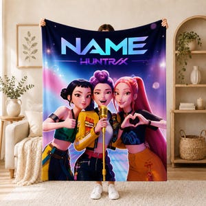 May include: A rectangular fleece blanket featuring a vibrant graphic of three animated characters standing together. The center character holds a gold microphone. The text "NAME" and "HUNTRAX" is printed in large, bold, blue letters at the top.