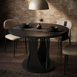 May include: A round black dining table with a central pedestal base, accompanied by two beige upholstered chairs. The table is set with a dark runner, two black bowls, and a small black vase containing a single dark flower in the center.