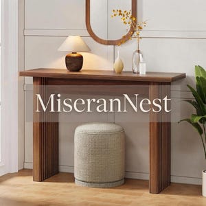 May include: A dark wood console table with vertical slat legs and a rectangular top. A beige fabric ottoman sits underneath. On the table are a lamp with a textured base, a glass vase with branches, and a small white vase. Text reads MiseranNest.