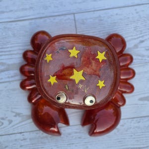 May include: A glossy, translucent orange resin crab figurine featuring a flat, circular top embedded with five bright yellow star shapes and a reddish-orange fish silhouette. The crab has two protruding googly eyes and eight rounded legs.