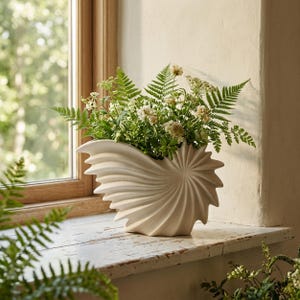 Puede incluir: Un jarrón de cerámica color crema con forma de concha de nautilo y una textura estriada en espiral. El jarrón está lleno de un exuberante arreglo de helechos verdes y pequeñas flores silvestres blancas, creando una composición natural.