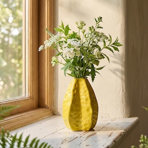 May include: A bright yellow ceramic vase with a textured, dimpled surface holding a bouquet of white wildflowers and green foliage. The vase has a tapered, multi-faceted shape and sits upright, showcasing a glossy finish that reflects light.