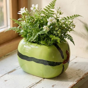 May include: A glossy, light green ceramic planter shaped like a rounded fruit, featuring dark green painted stripes and a small brown stem detail. The planter is filled with a lush arrangement of green fern fronds and small white flowers.