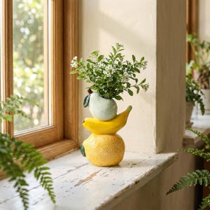 May include: A ceramic vase shaped like a stack of fruit, featuring a textured yellow lemon at the base, a bright yellow banana in the middle, and a pale green pear on top, holding a bouquet of small white flowers with green leaves.