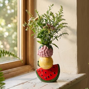 Può includere: Un vaso in ceramica a forma di pila di frutta, con una base a fetta di anguria rossa, un limone giallo al centro e un lampone rosa in cima. Il vaso contiene un bouquet di steli verdi e piccoli fiori di campo, con una finitura lucida.