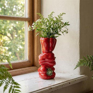 Puede incluir: Un jarrón de cerámica roja brillante con forma de pila de tres pimientos. El jarrón tiene una superficie lisa y reflectante y contiene un ramo de pequeñas flores blancas con tallos y hojas verdes, colocado en posición vertical.