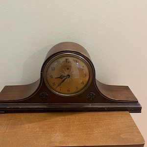 Puede incluir: Un reloj de sobremesa de madera marrón con una parte superior redondeada y una base ancha. La esfera es circular con un borde dorado, números arábigos negros y manecillas negras. La palabra Telechron está impresa en el centro sobre las manecillas.