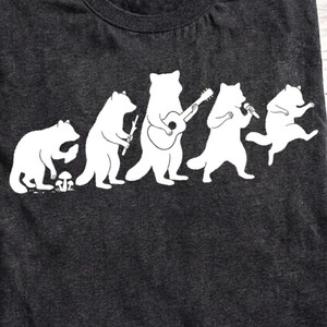 May include: A dark charcoal grey crew neck t-shirt featuring a white graphic print of five raccoons in a row. The raccoons are depicted in various poses, including one foraging near a mushroom, one holding a stick, one playing a guitar, one singing into a microphone, and one dancing.