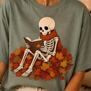 Peut inclure: Un t-shirt vert sauge avec une illustration graphique d'un squelette portant une écharpe rouille, assis dans un tas de feuilles d'automne colorées tout en lisant un livre marron avec un motif de feuille d'érable dorée sur la couverture.