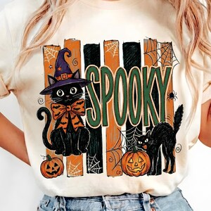 Peut inclure: Un t-shirt crème avec une impression graphique représentant deux chats noirs, l'un portant un chapeau de sorcière violet et un nœud orange, l'autre près d'une citrouille. Le motif inclut le mot SPOOKY en vert, des rayures orange et noires, et des toiles d'araignée.