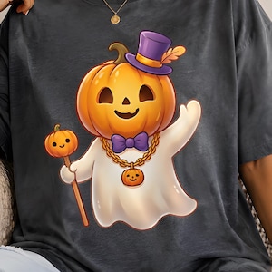 Peut inclure: Un t-shirt gris anthracite orné d'un motif représentant un fantôme à tête de citrouille, portant un haut-de-forme violet, un nœud papillon violet et une chaîne dorée avec un petit pendentif citrouille. Le fantôme tient une citrouille sur un bâton.