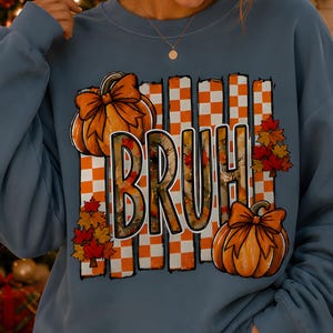 Peut inclure: Un sweat-shirt bleu cendré à col rond arborant un motif central avec le texte "BRUH" en lettres grasses. Le design comprend un fond à carreaux orange et blanc, deux citrouilles orange avec des nœuds et des grappes de feuilles d'automne.