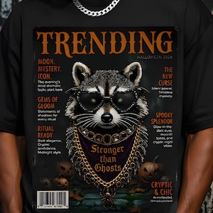 May include: A black t-shirt featuring a graphic print of a raccoon wearing heart-shaped sunglasses and a gold chain with a spider pendant. The print resembles a magazine cover titled TRENDING HALLOWEEN 2026 with text including Stronger than Ghosts.