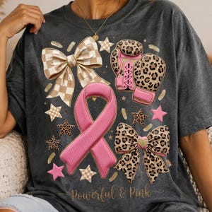 May include: A charcoal gray t-shirt featuring a graphic print of a pink awareness ribbon, two leopard print bows, and a pair of leopard print boxing gloves. The design includes small stars and the gold cursive text "Powerful & Pink" at the bottom.