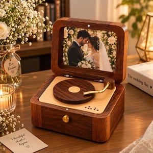 Op de afbeelding: Een houten muziekdoos ontworpen als een platenspeler met een donkerhouten frame en een lichthouten basis. Het deksel bevat een foto van een koppel in trouwkleding. Een goudkleurige toonarm rust op een ronde houten schijf en een gouden knop voorop.