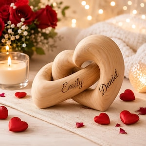 May include: Two interlocking light-colored wooden heart sculptures featuring the engraved names Emily and Daniel. The smooth, curved wooden hearts are positioned together, showcasing a natural wood grain texture and dark, cursive lettering.