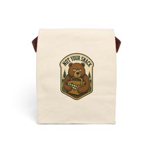 May include: A cream-colored canvas lunch bag featuring a graphic illustration of a brown bear holding a picnic basket. The bear has an annoyed expression, and the text above it reads NOT YOUR SNACK. The design is enclosed in a shield-shaped border.