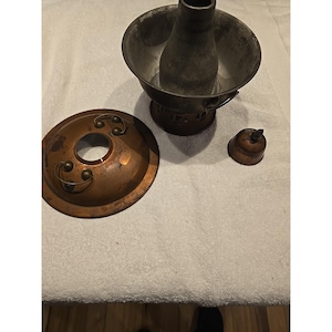 May include: A copper-colored metal hot pot with a central chimney, a separate circular lid featuring two small ring handles, and a small, pointed cap. The main vessel has a dark, weathered interior and a perforated base for heat circulation.