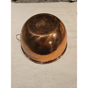 May include: A round, polished copper mixing bowl with a smooth, reflective surface and a warm, metallic orange-brown tone. The bowl features a small, circular brass ring handle attached to one side, designed for hanging or easy handling during use.