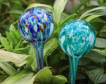 Könnte beinhalten: Zwei kugelförmige Pflanzenbewässerungskugeln aus Glas mit langen Stielen. Die linke Kugel zeigt ein gesprenkeltes Muster aus blauem, weißem und bernsteinfarbenem Glas. Die rechte Kugel hat ein gesprenkeltes Muster aus blaugrünem und weißem Glas.