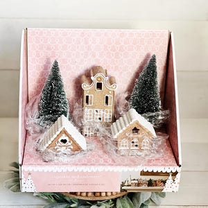 May include: A set of five LED holiday decorations in a pink box, including two green bottle brush trees, one tall brown house with white trim, and two smaller brown houses with white roofs. The box features the text cupcakes and cashmere.