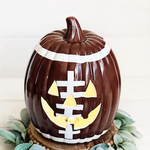 May include: A brown ceramic pumpkin shaped like a football with white painted laces and stripes. The pumpkin features a carved jack-o'-lantern face with triangular eyes and a jagged mouth, illuminated from within by a warm light.