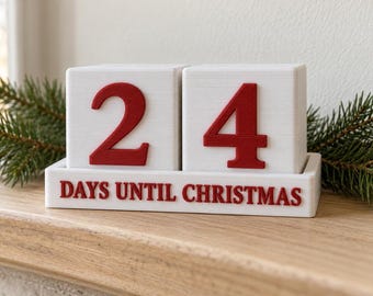 May include: A white block countdown calendar featuring two large cubes with red numbers and a base tray with the red text "DAYS UNTIL CHRISTMAS" printed on the front. The cubes currently display the number 24, set against a festive evergreen branch.